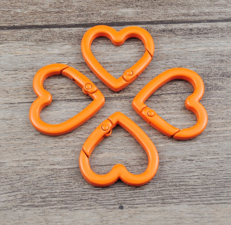 May include: Four orange heart-shaped carabiner clips arranged in a cloverleaf pattern. Each clip has a spring-loaded gate for secure attachment. The clips are made of a smooth, solid material and are set against a wooden background.