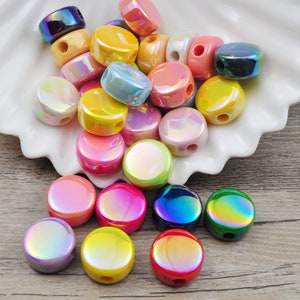 May include: A collection of iridescent round beads in various colors, including pink, yellow, blue, green, and purple. The beads have a hole through the center and are arranged in a pile on a white surface.