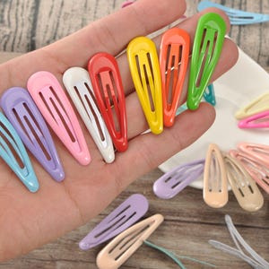 May include: A collection of colorful, oval-shaped hair clips in various shades, including blue, purple, pink, white, red, yellow, orange, and green. Some clips are held in a hand, while others are scattered on a white plate and surface.