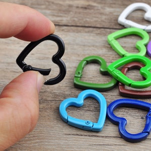 May include: A hand holding a black heart-shaped carabiner clip, with several other colorful heart-shaped clips in the background. The clips are in various colors, including green, blue, and red. The clips are made of metal.
