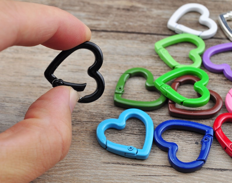 May include: A hand holding a black heart-shaped carabiner clip, with several other colorful heart-shaped clips in the background. The clips are in various colors, including green, blue, and red. The clips are made of metal.