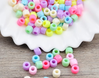 Puede incluir: Un conjunto de cuentas de plástico de colores pastel, incluyendo rosa, amarillo, verde, azul y morado. Las cuentas cilíndricas tienen un agujero en el centro, ideales para bisutería o manualidades. Algunas cuentas están dispersas sobre una superficie de madera.