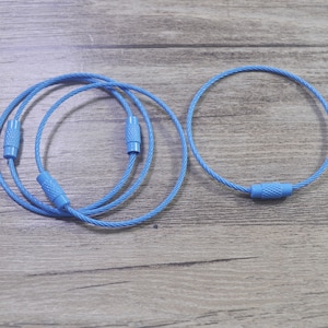 Puede incluir: Varios llaveros azul claro se muestran sobre un fondo de madera. Cada llavero es un bucle circular de cable de acero trenzado, asegurado con un cierre de tornillo azul a juego. Los llaveros están diseñados para sujetar llaves u otros objetos pequeños.