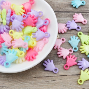 Op de afbeelding: Een kom gevuld met kleurrijke plastic handcharms. De charms zijn in verschillende pastelkleuren, waaronder roze, blauw, geel en paars. Elke charm heeft een klein gaatje om aan een armband of ketting te bevestigen.
