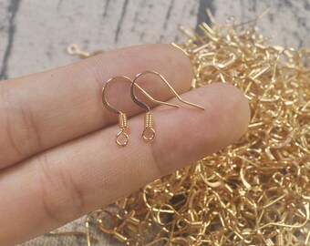 May include: Two gold-toned earring hooks, also known as ear wires, are shown on a hand. The hooks are made of metal and have a spring-loaded closure. The hooks are commonly used for making earrings.