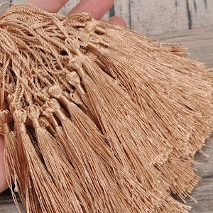 May include: A collection of numerous champagne-coloured silk tassels, each featuring a tightly knotted head and a long, flowing fringe. The tassels are connected by thin, twisted cords, creating a dense, uniform cluster of shimmering, light-reflecting threads.