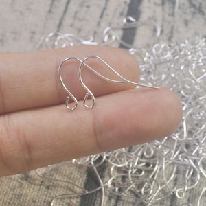 May include: Silver earring hooks, a close-up of two earring hooks held in a hand, with a pile of earring hooks in the background.