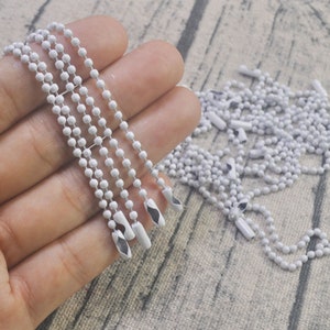 May include: A close-up of a white metal chain with a silver finish. The chain is made of small, round beads and has a loop at the end. The chain is lying on a white surface.