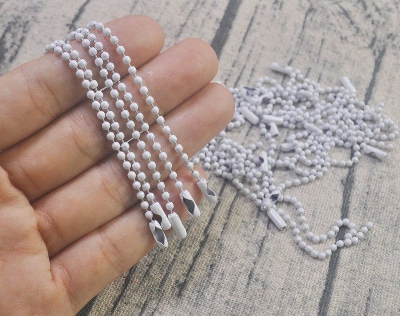 May include: A close-up of a white metal chain with a silver finish. The chain is made of small, round beads and has a loop at the end. The chain is lying on a white surface.