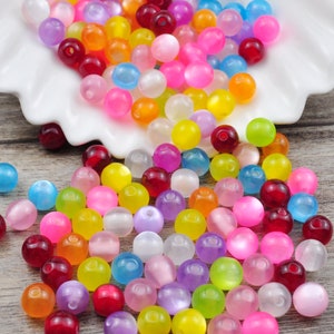 Può includere: Un vasto assortimento di perline rotonde colorate. Le perline sono in plastica e hanno una finitura traslucida e iridescente. Le perline sono sparse su una superficie bianca.