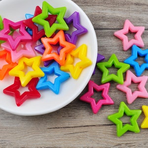 May include: A collection of colorful plastic star-shaped beads in various colors, including red, orange, yellow, green, blue, purple, and pink. The beads are arranged in a pile on a white plate and a wooden surface.