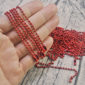 May include: A close-up of a hand holding several strands of red metal chain. The chain is made of small, round beads and has a shiny finish. The chain is lying on a brown, textured surface.