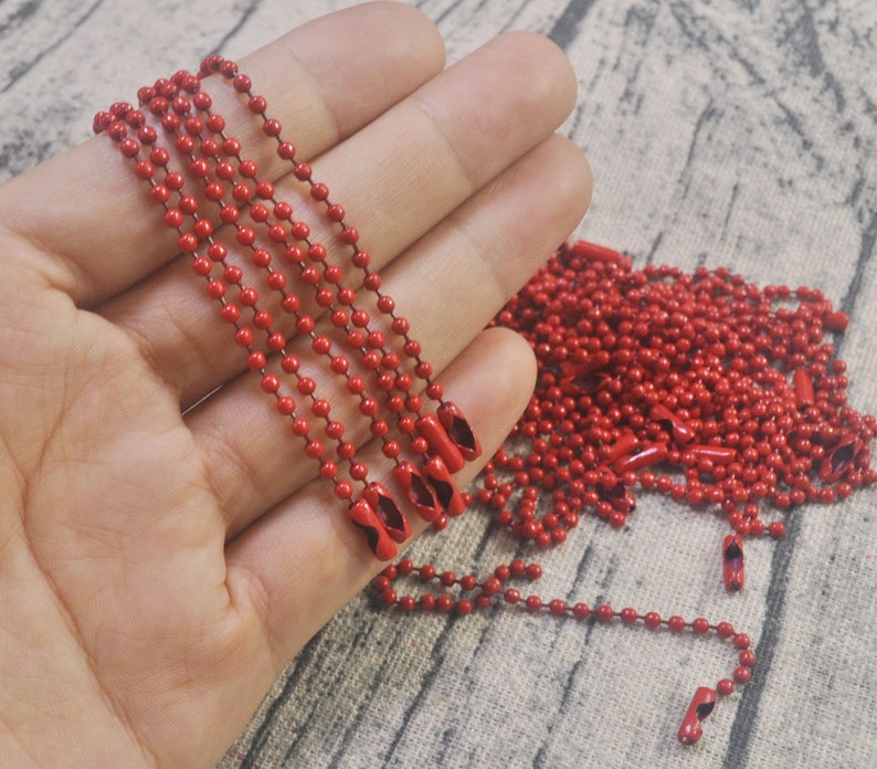 May include: A close-up of a hand holding several strands of red metal chain. The chain is made of small, round beads and has a shiny finish. The chain is lying on a brown, textured surface.