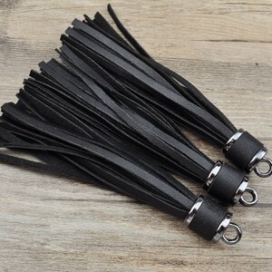 Può includere: Tre nappine in pelle nera con anelli in metallo argentato. Le nappine sono fatte di sottili strisce di pelle e sono lunghe circa 15 cm.