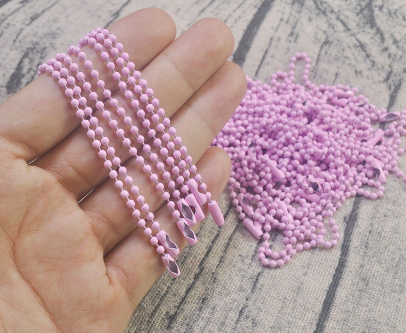 May include: A close-up of a pile of pink metal chains. The chains are made of small, round links and have a shiny finish. The chains are arranged in a loose pile, with some of the chains hanging over the edge of the pile.