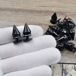 May include: Two black metal pyramid-shaped studs with a silver ring around the base. The studs are on a white surface. There are several more studs in the background.