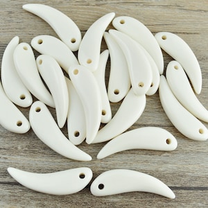 May include: A collection of white, tooth-shaped pendants with a hole at the top for stringing. The pendants are made of a smooth, polished material and have a slightly curved shape.