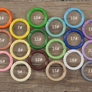 Puede incluir: Una colección de cierres metálicos redondos y coloridos en varios tonos, incluyendo rojo, naranja y verde. Cada cierre está numerado y tiene una abertura con bisagras. Los cierres están dispuestos sobre una superficie de madera.
