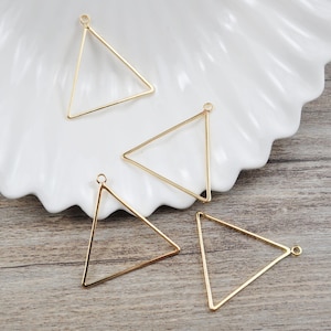 Puede incluir: Cuatro marcos metálicos de forma triangular de color dorado con un pequeño bucle en la parte superior. Los marcos están sobre una superficie blanca y marrón.