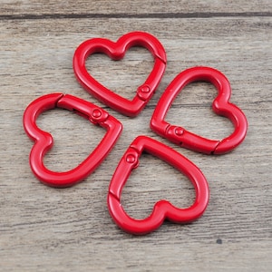 May include: Four red heart-shaped carabiner clips. Each clip has a spring-loaded gate for secure closure. The clips are made of a shiny, solid red material and are arranged on a wooden surface.