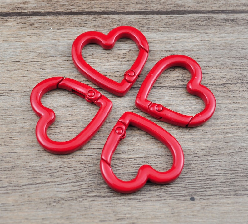 May include: Four red heart-shaped carabiner clips. Each clip has a spring-loaded gate for secure closure. The clips are made of a shiny, solid red material and are arranged on a wooden surface.