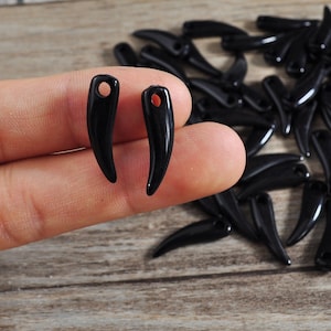 May include: Two black horn-shaped pendants with a hole at the top for stringing. The pendants are on a finger, and there are many more similar pendants in the background.