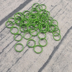 May include: A collection of small, bright green metal rings scattered on a textured, light gray surface. The rings are evenly coated with a glossy finish. These rings are likely used for crafting or jewelry making.