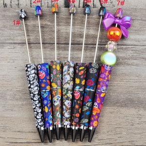 Könnte beinhalten: Sieben dekorative Stifte mit verschiedenen Designs. Die Stifte haben Schädel-, Blumen- und Dia de los Muertos-Muster. Die Stiftspitzen umfassen einen Totenkopf, bunte Perlen und eine lila Schleife. Die Stifte sind zum Schreiben.
