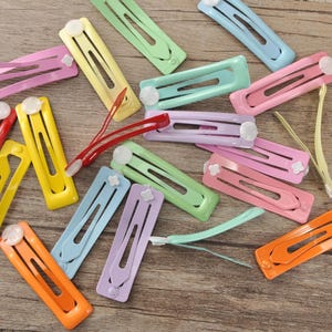 May include: A collection of colorful hair barrettes in various shades, including pink, yellow, green, and blue. Each barrette features a decorative white flower accent. The barrettes are scattered on a wooden surface.