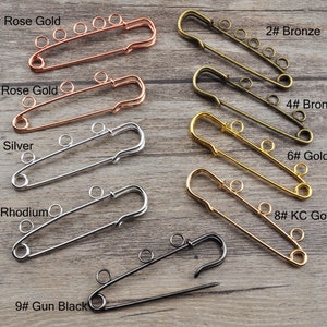 Pode incluir: Nove alfinetes de segurança em diferentes acabamentos, incluindo ouro rosa, bronze, prata, ródio e ouro. Os alfinetes estão dispostos em duas filas sobre uma superfície de madeira. Os alfinetes estão numerados de 1 a 9.