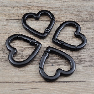 May include: Four black heart-shaped carabiner clips are arranged on a wooden surface. The clips are made of metal and have a spring-loaded gate for easy attachment. These decorative clips can be used for keychains, jewelry, or other craft projects.