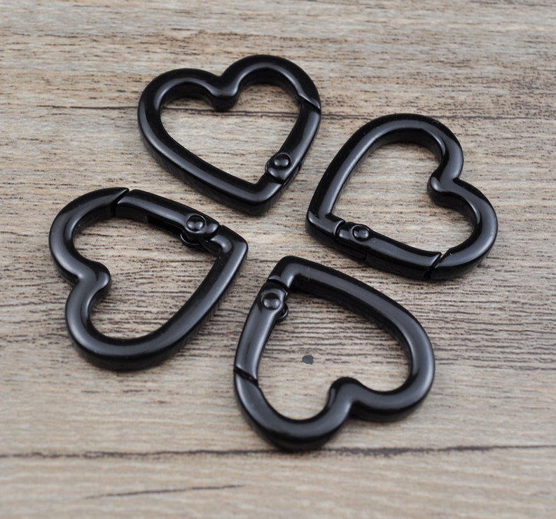 May include: Four black heart-shaped carabiner clips are arranged on a wooden surface. The clips are made of metal and have a spring-loaded gate for easy attachment. These decorative clips can be used for keychains, jewelry, or other craft projects.