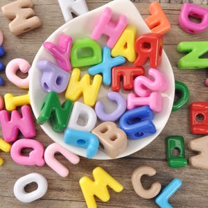 May include: A collection of colorful alphabet beads in various hues, including pink, yellow, green, and blue. The beads are scattered on a wooden surface, with some contained in a white, shallow dish. These plastic beads are ideal for crafting and jewelry making.
