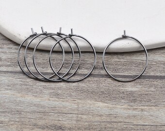 May include: Several silver-colored metal hoop earring findings are arranged on a light-colored wooden surface. The hoops are circular with a small loop at the top for attaching to an earring hook. The background is a white, curved surface.