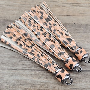 May include: Three brown and black leopard print tassel keychains with black metal rings. The tassels are made of leather and have a fringe design.