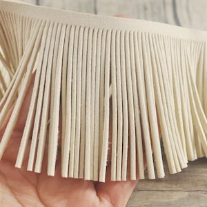 Puede incluir: Primer plano de una tira de flecos de cuero blanco roto. Los flecos tienen aproximadamente 2,5 cm de ancho y una textura suave y similar al ante.