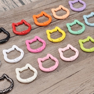 May include: A collection of colorful cat-shaped metal clasps in various colors, including black, red, orange, pink, yellow, green, blue, and silver. The clasps are arranged on a wooden surface.