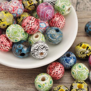 May include: A collection of colorful wooden beads with intricate patterns and designs. The beads are in various colors, including red, blue, green, yellow, and pink, and are displayed in a white bowl and scattered on a wooden surface. These beads are ideal for jewelry making.