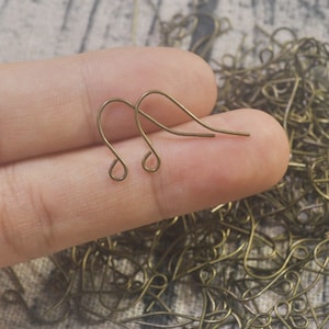May include: Two gold-toned earring hooks, also known as French hooks, are shown in a close-up image. The hooks are made of metal and have a simple, elegant design.
