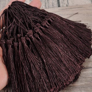 Peut inclure: Une collection de pompons décoratifs brun foncé, chacun doté d'une boucle supérieure nouée et d'une grappe dense de fils fins et soyeux suspendus. Les pompons sont uniformes en couleur et en texture, affichant une teinte brune riche et profonde.
