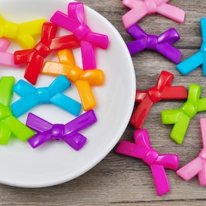 Puede incluir: Una colección de botones de plástico de colores en forma de lazo en varios colores, incluyendo rojo, amarillo, azul, verde, morado y rosa. Los botones están dispuestos en un plato blanco y una superficie de madera.