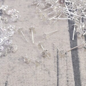 May include: A close-up of silver earring findings, including post and backs, on a textured fabric surface.