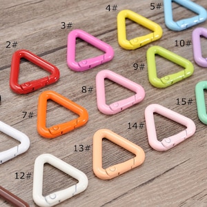 May include: A selection of sixteen triangular carabiner clips in a range of colours, including black, red, pink, yellow, blue, purple, green, orange, and beige. Each clip is approx. 2.5 cm wide and has a spring-loaded gate for secure attachment.