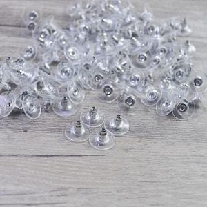 Puede incluir: Un montón grande de respaldos de pendientes de plástico transparente con postes de metal plateado. Hay algunos respaldos de pendientes esparcidos sobre la superficie de madera.
