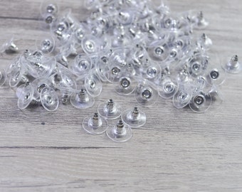 May include: A large pile of clear plastic earring backs with silver metal posts. There are a few earring backs scattered on the wooden surface.