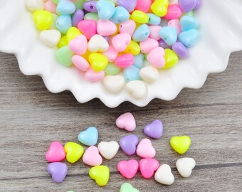 May include: A bowl filled with colorful heart-shaped beads in shades of pink, purple, yellow, and green. The beads are scattered on a wooden surface.