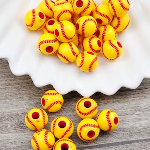 Pode incluir: Um monte de contas amarelas e vermelhas em forma de bola de softball com um furo no centro. As contas estão em um prato branco e uma superfície de madeira.