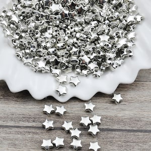 May include: A white bowl filled with silver star-shaped beads. The beads are scattered on a wooden surface.