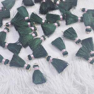 Puede incluir: Una colección de borlas de color verde oscuro con ribete rosa y anillos dorados. Las borlas están esparcidas sobre una superficie blanca y peluda.