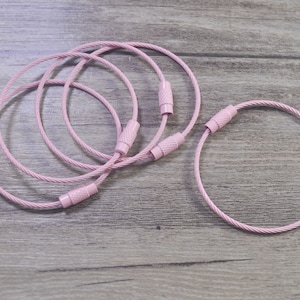 Peut inclure: Cinq porte-clés en câble métallique rose avec un design de câble tressé. Les porte-clés sont empilés les uns sur les autres.